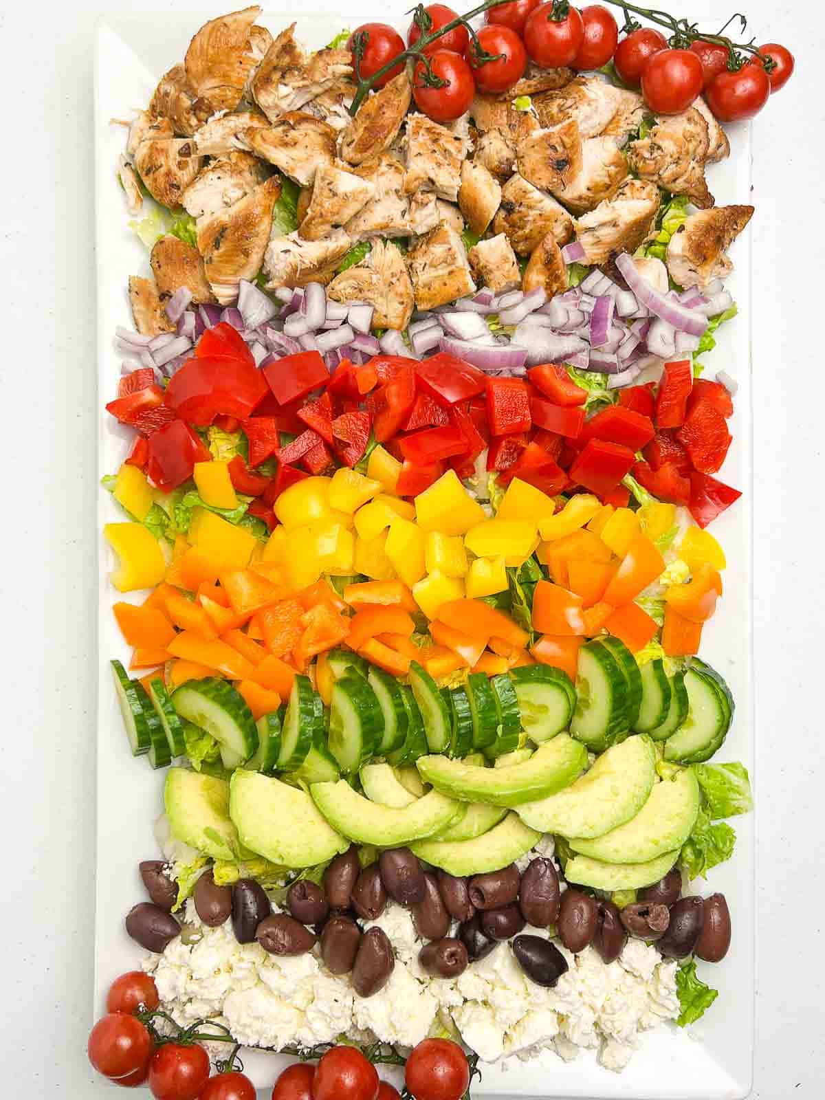 Overhead view of a large white rectangular platter with Greek Chicken Cobb Salad ingredients in rows.