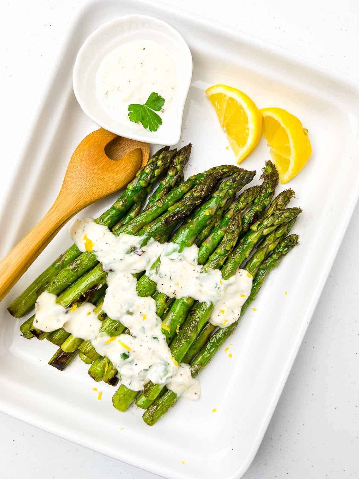 Close up view of asparagus spears on a white platter with lemon garlic aioli and lemon slices.