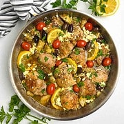A skillet with baked lemon artichoke chicken thighs, topped with lemon slices, olives, tomatoes and parsley.