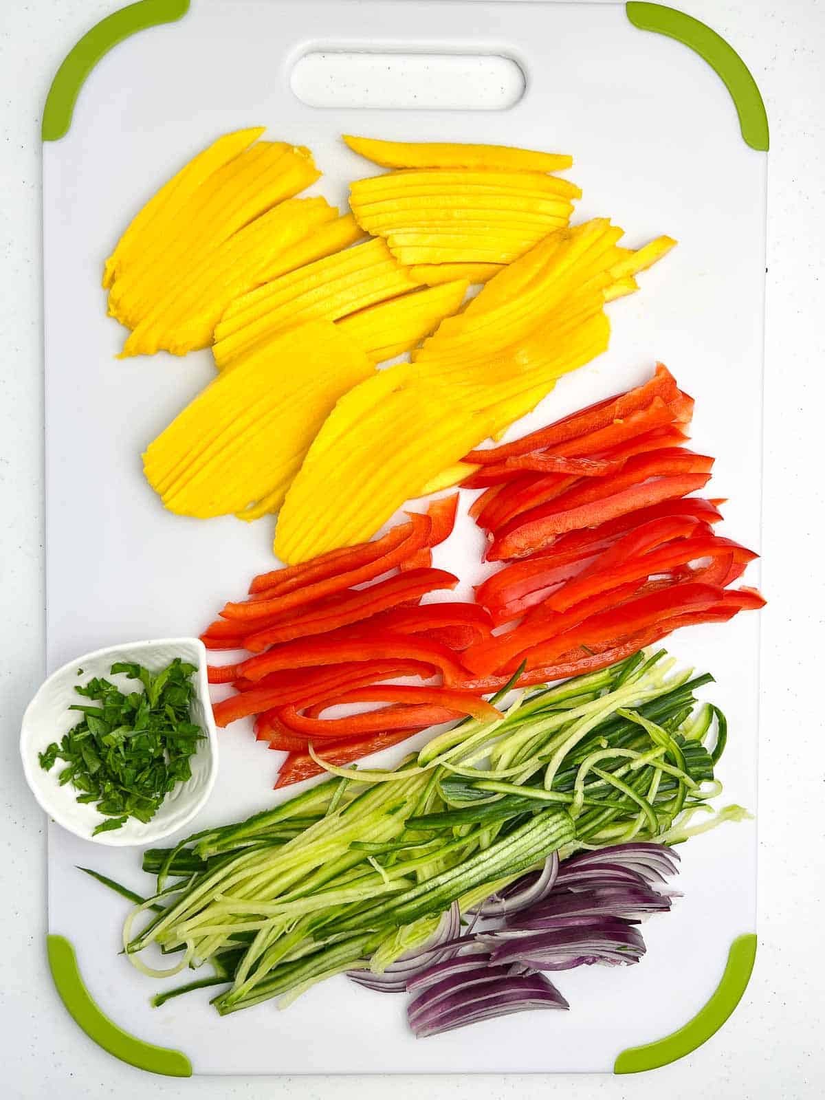 Sliced mango, red bell pepper, cucumber, red onion and cilantro on a cutting board.