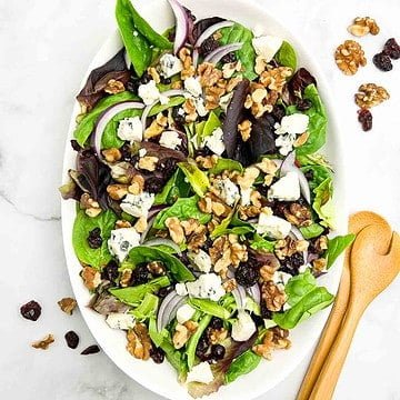 Overhead view of an oval bowl with blue cheese and walnut salad with a pair of tongs beside.