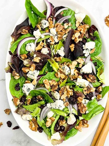 Overhead view of an oval bowl with blue cheese and walnut salad with a pair of tongs beside.