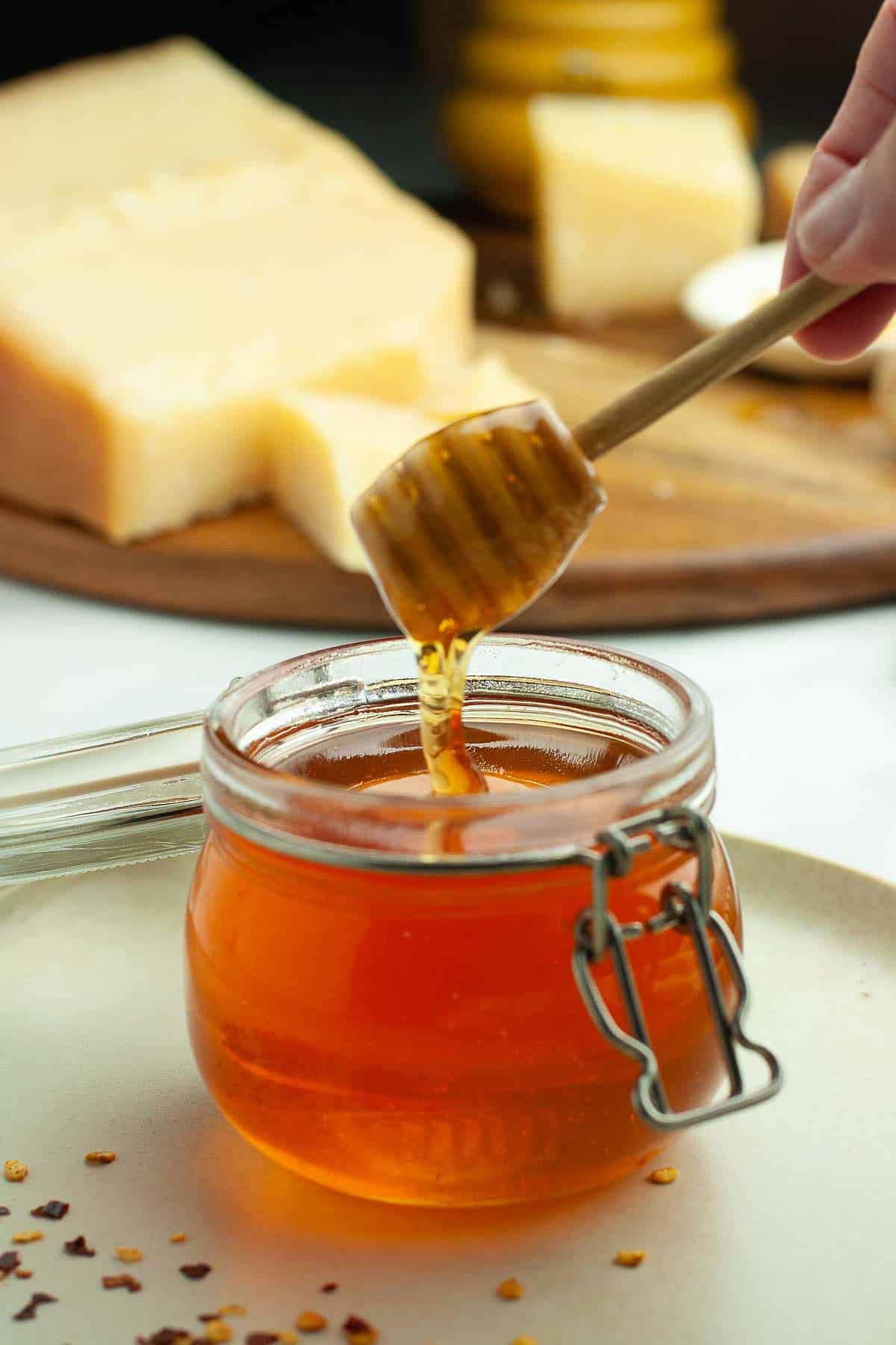 Dipping a honey dipper into a small jar of hot honey with Parmesan cheese in the background.
