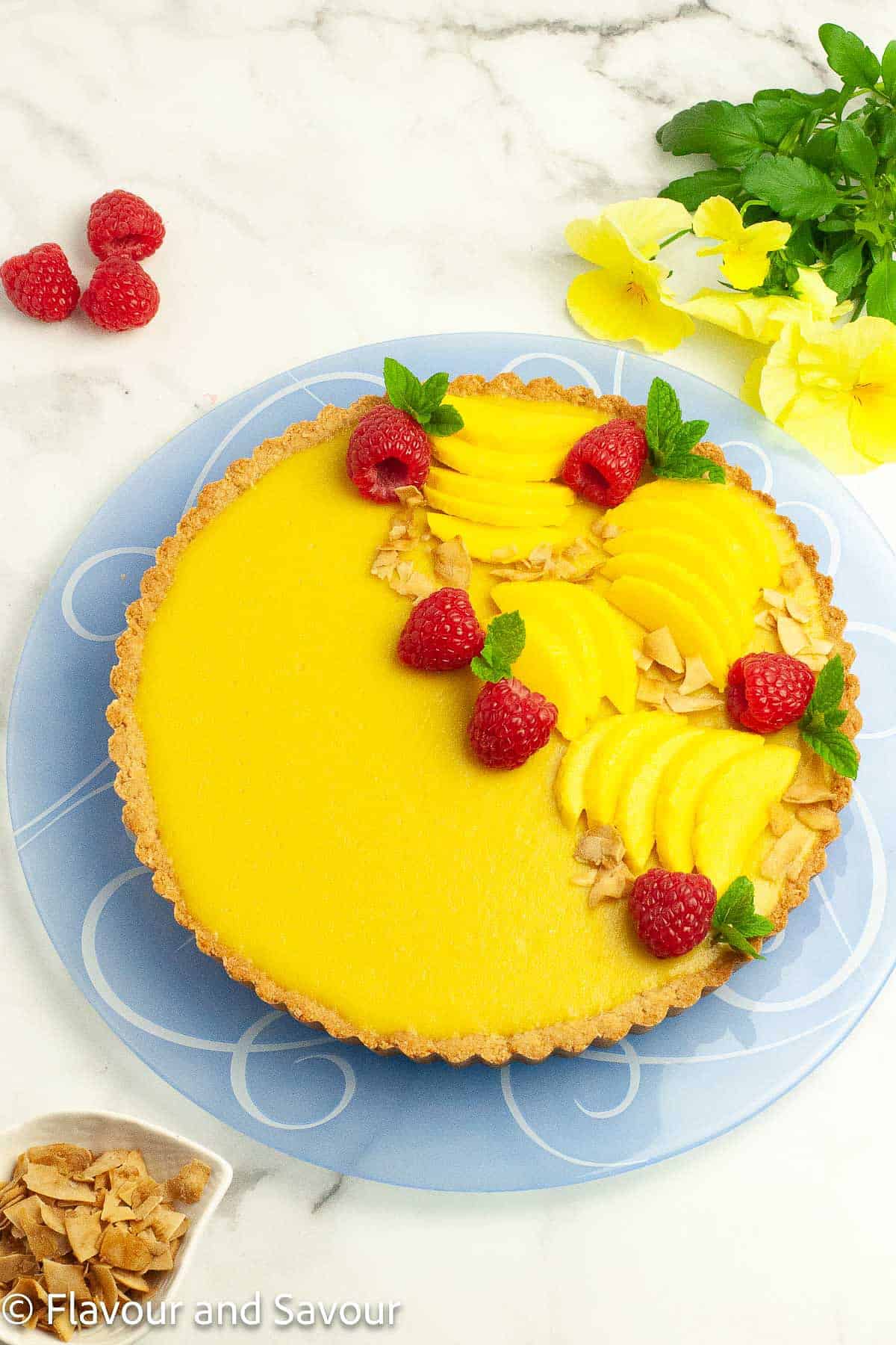 Overhead view of a gluten-free mango tart garnished with sliced mango, raspberries, and coconut chips.