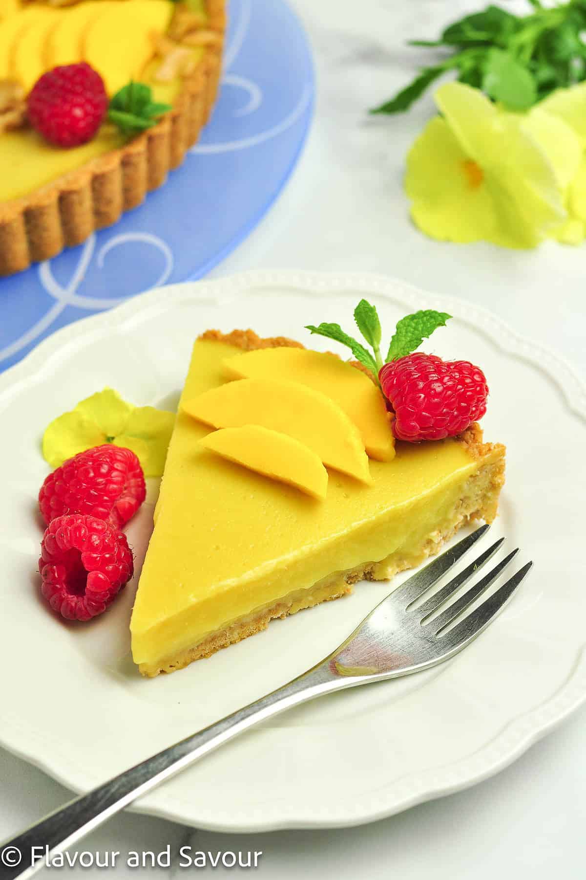 A slice of mango tart on a plate with raspberries and sliced mango.