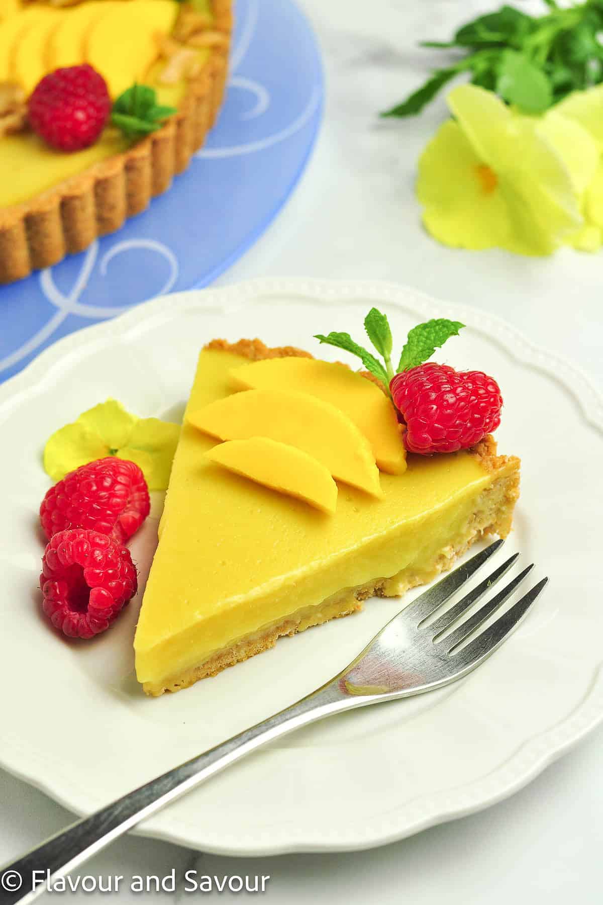 A slice of mango tart on a plate with raspberries and sliced mango.