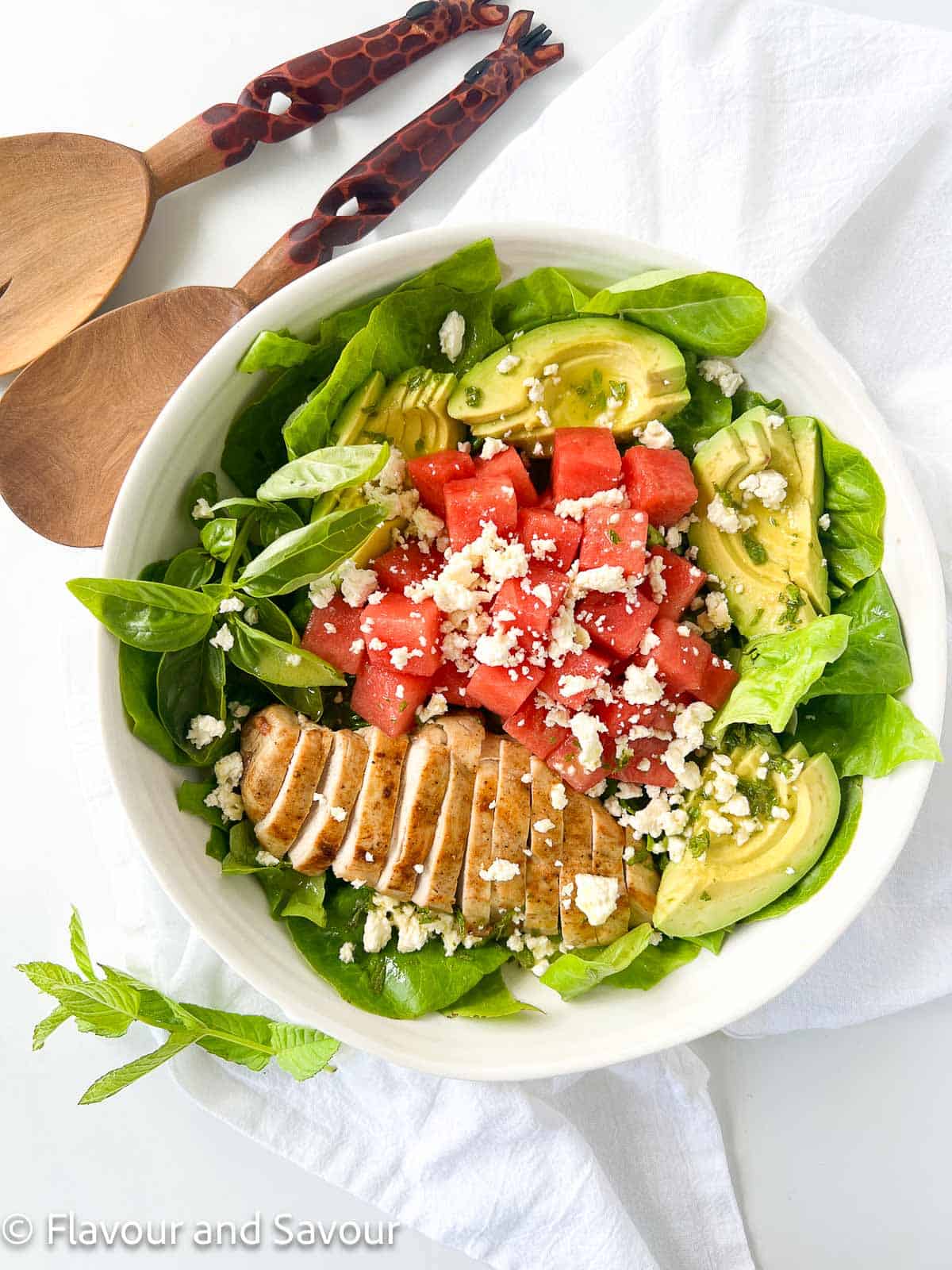 A bowl of greens with sliced chicken breast, watermelon cubes, avocado slices and feta cheese.