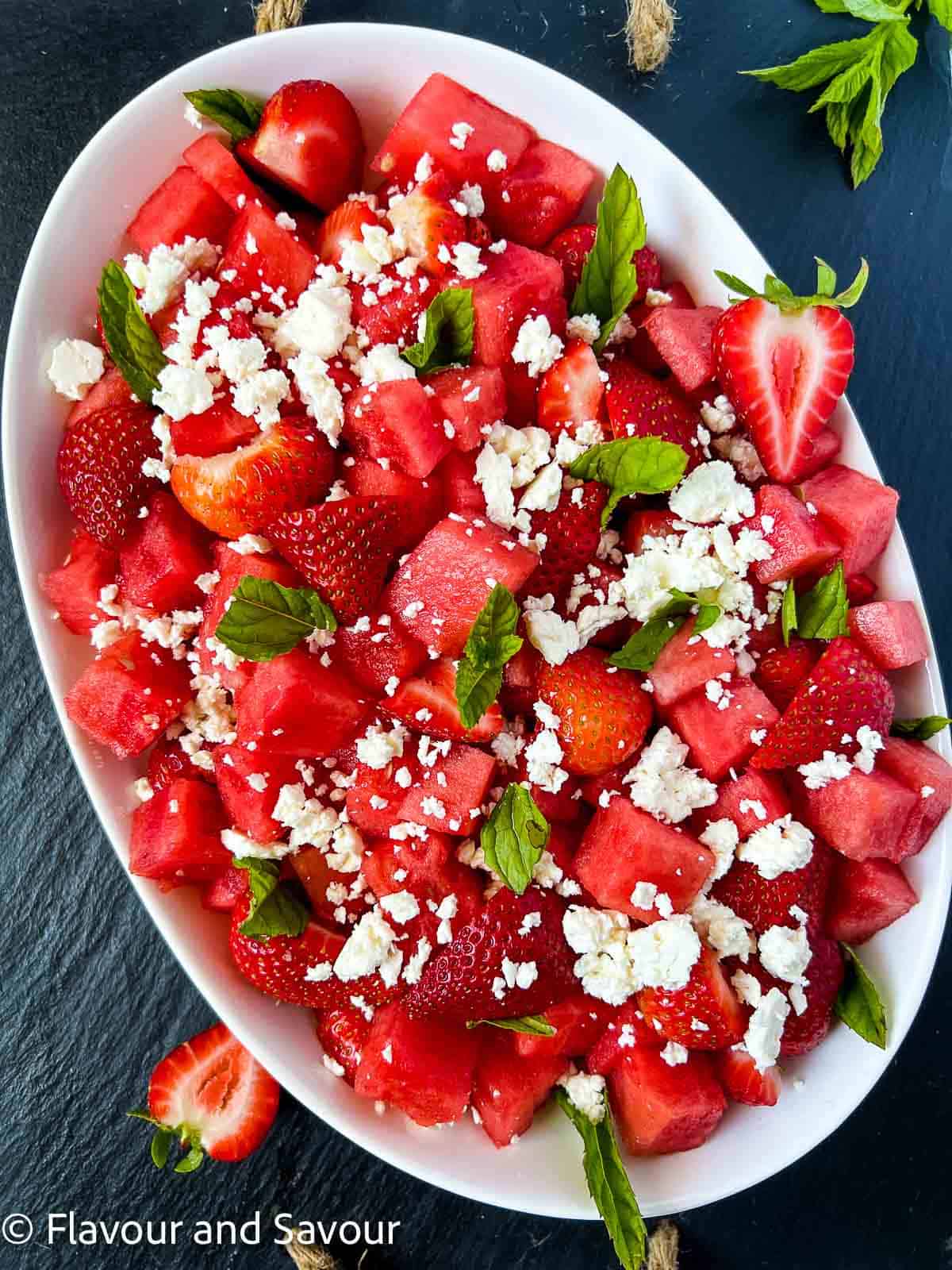 Overhead view of an oval bowl with strawberry watermelon salad with crumbled feta cheese and fresh mint leaves.