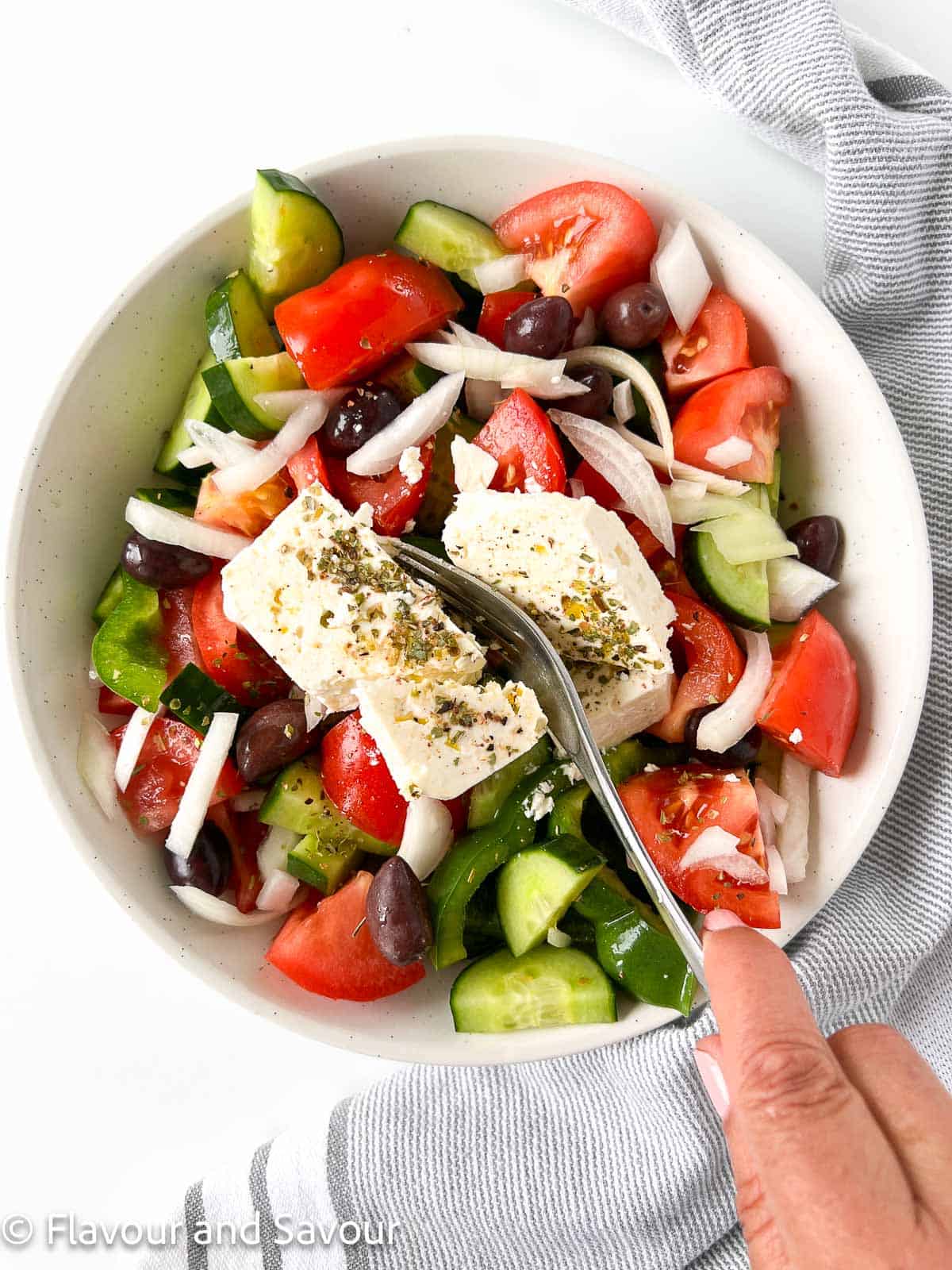 A Greek salad with a fork breaking apart a block of feta cheese on top.