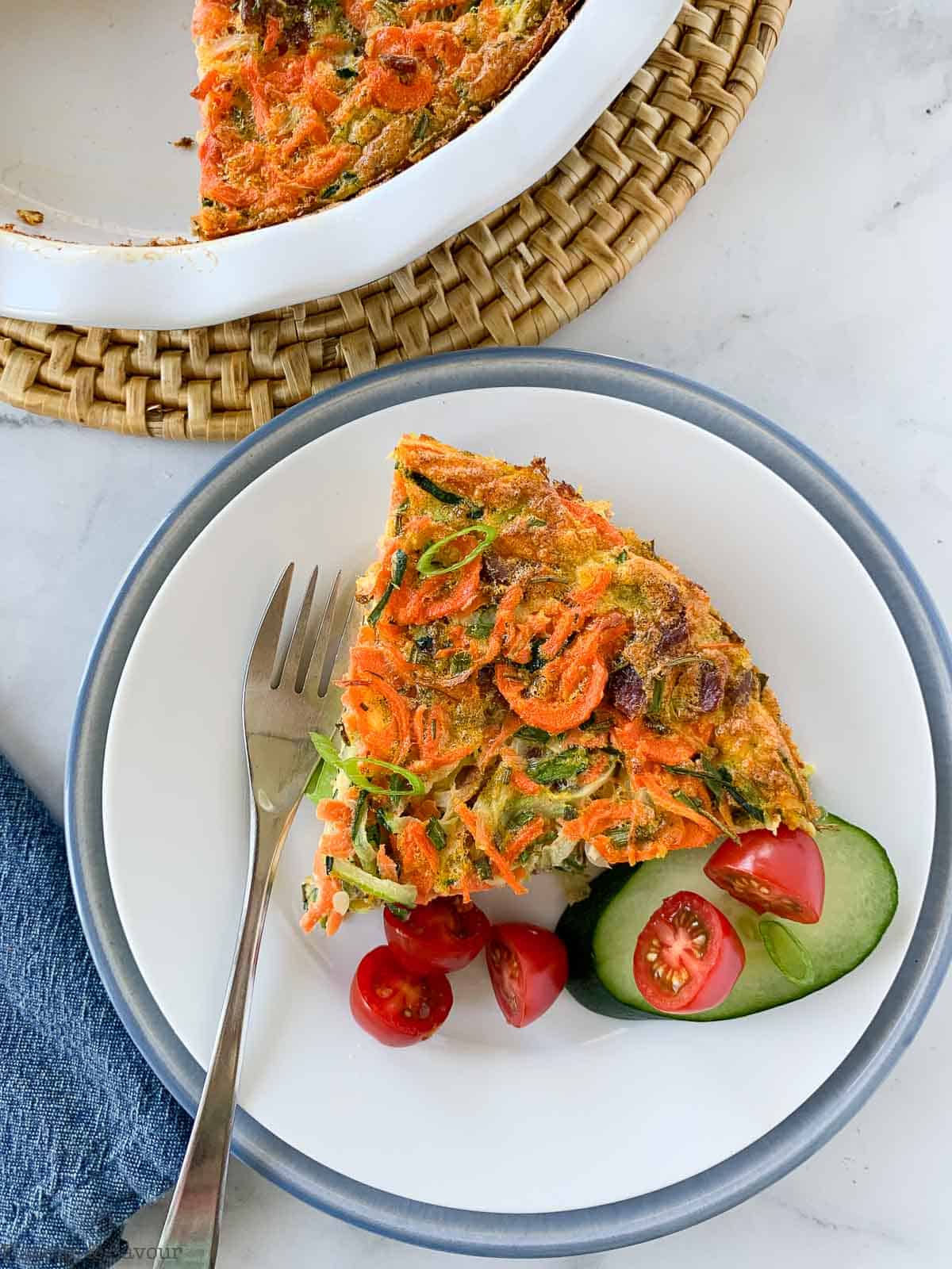 A wedge of zucchini carrot quiche on a plate with cucumber and tomatoes.