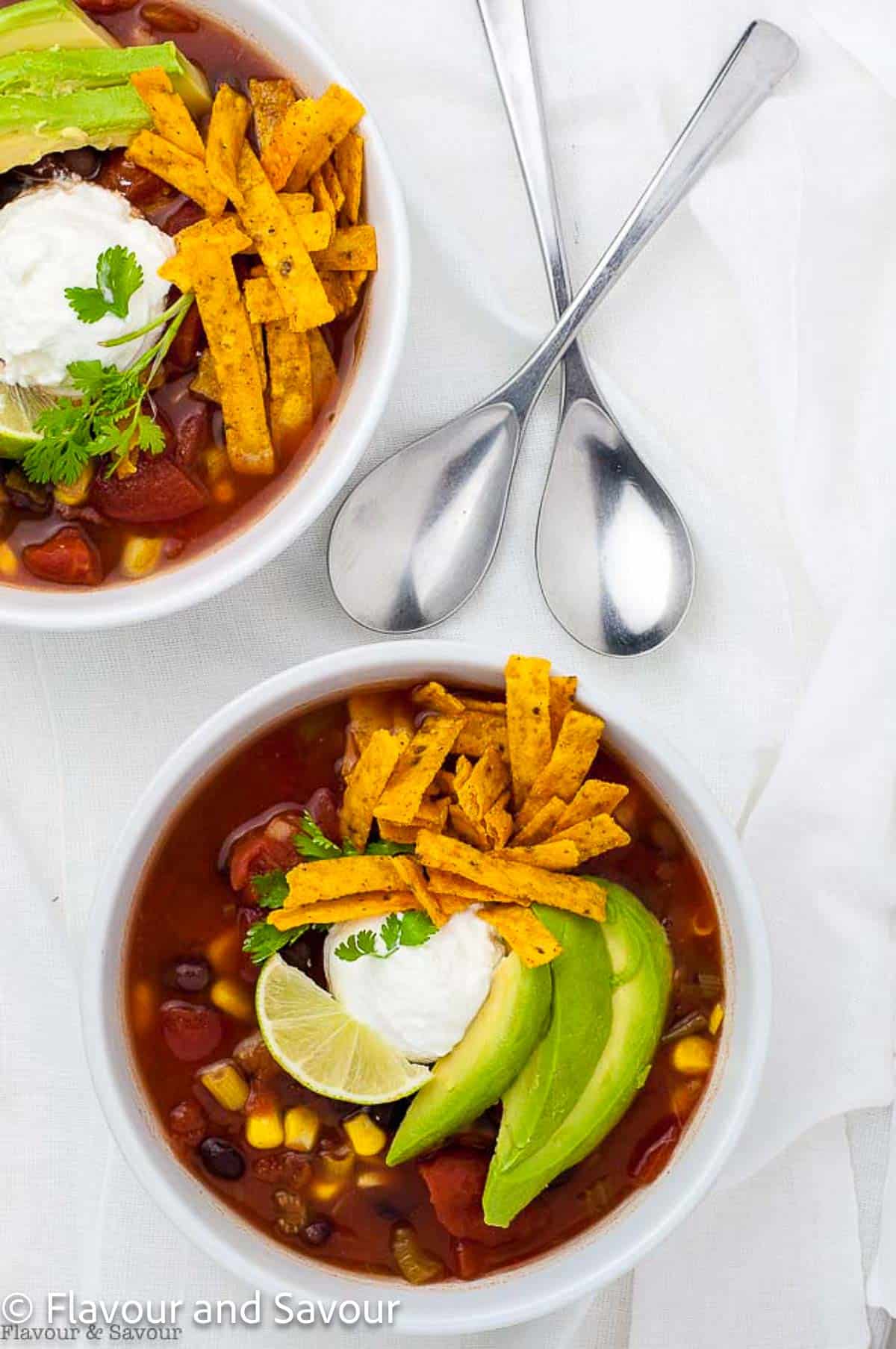Overhead view of two bowls of black bean soup cooked in a slow cooker and garnished with avocado and tortilla strips.