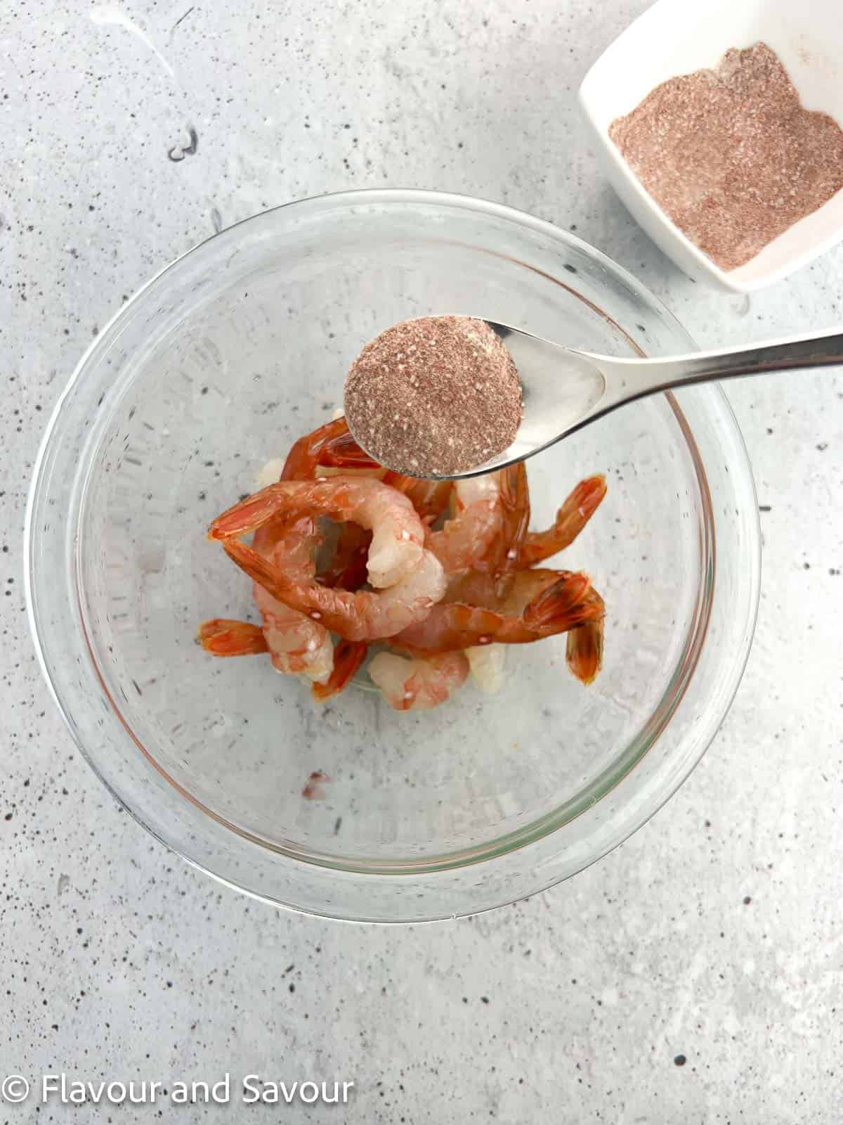 Adding a spoonful of shrimp seasoning mix to a bowl of raw spot prawns.