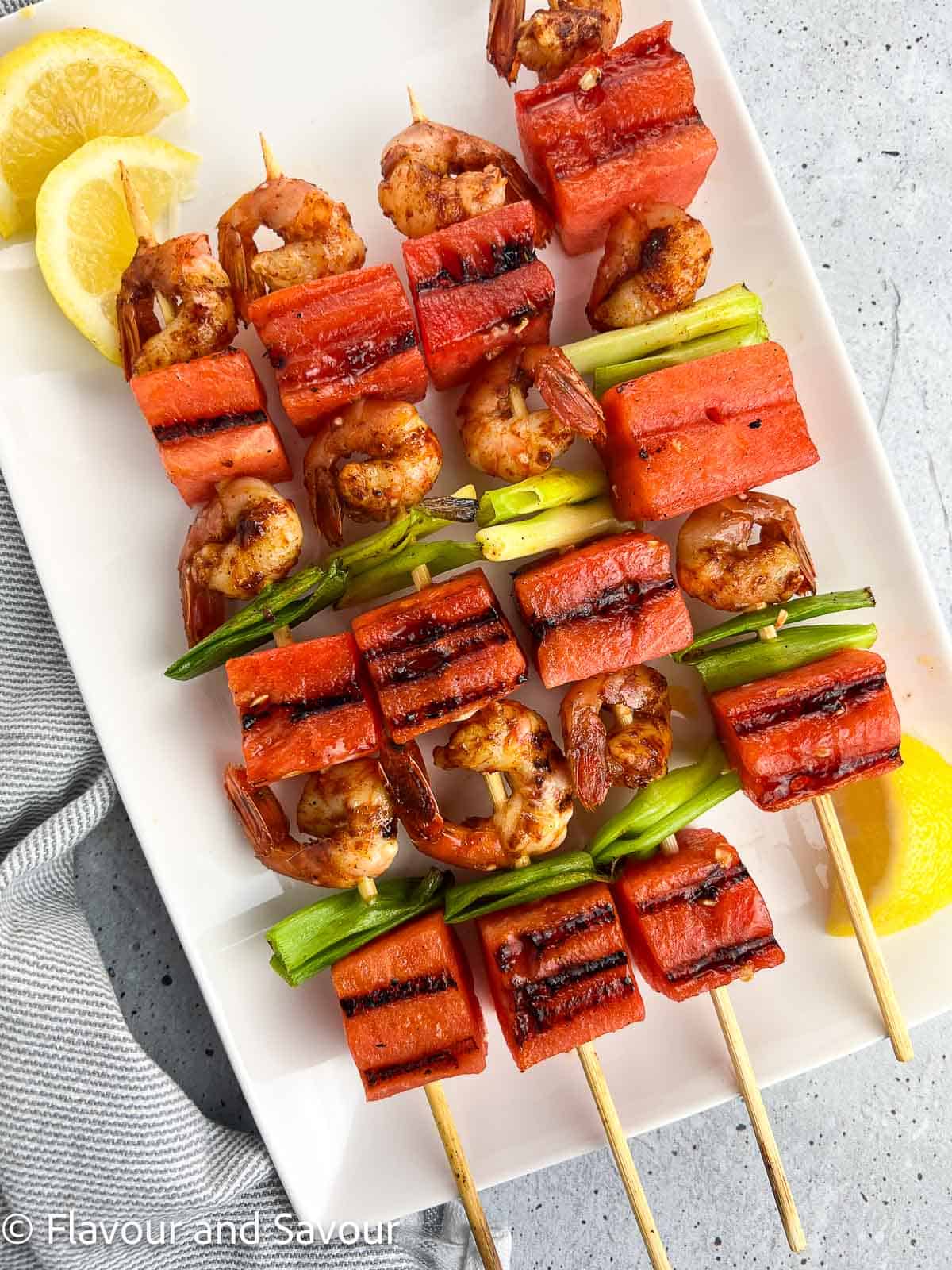 Spot prawn and watermelon kabobs on a white platter with lemon wedges.