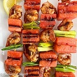 Square image of grilled watermelon spot prawn kabobs on a white platter.