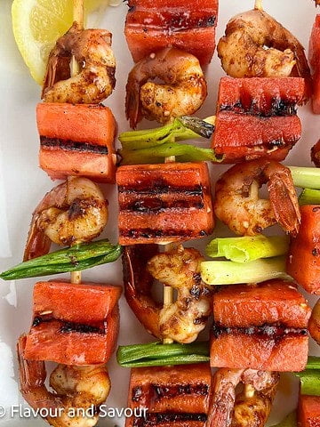 Square image of grilled watermelon spot prawn kabobs on a white platter.