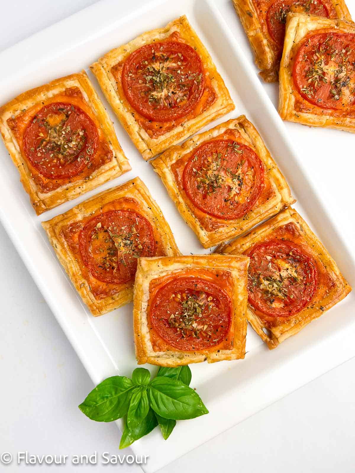 Puff pastry cheese and tomato tarts on a white serving plate with fresh basil leaves.