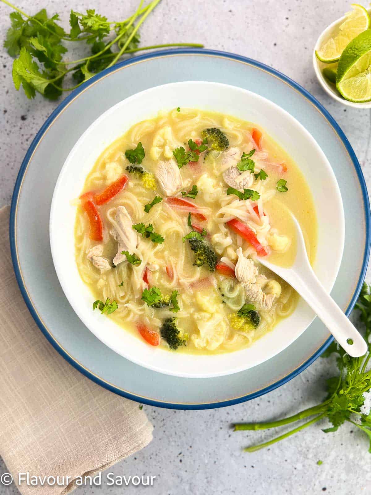 Overhead view of a bowl of Thai green curry chicken noodle soup with lime wedges beside it.