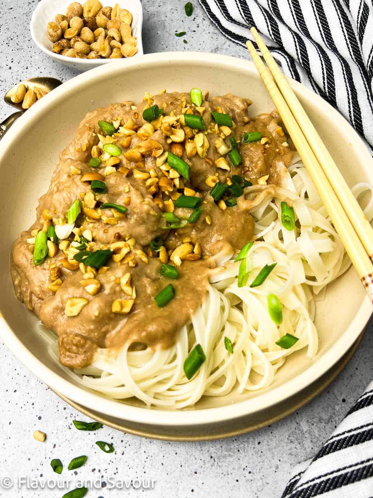 Thai peanut chicken thighs with rice noodles in a bowl, garnished with chopped salted peanuts and sliced green onions.