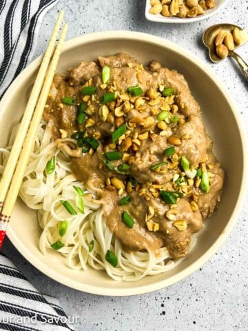 A shallow bowl with Thai peanut chicken and noodles garnished with chopped peanuts and green onions.