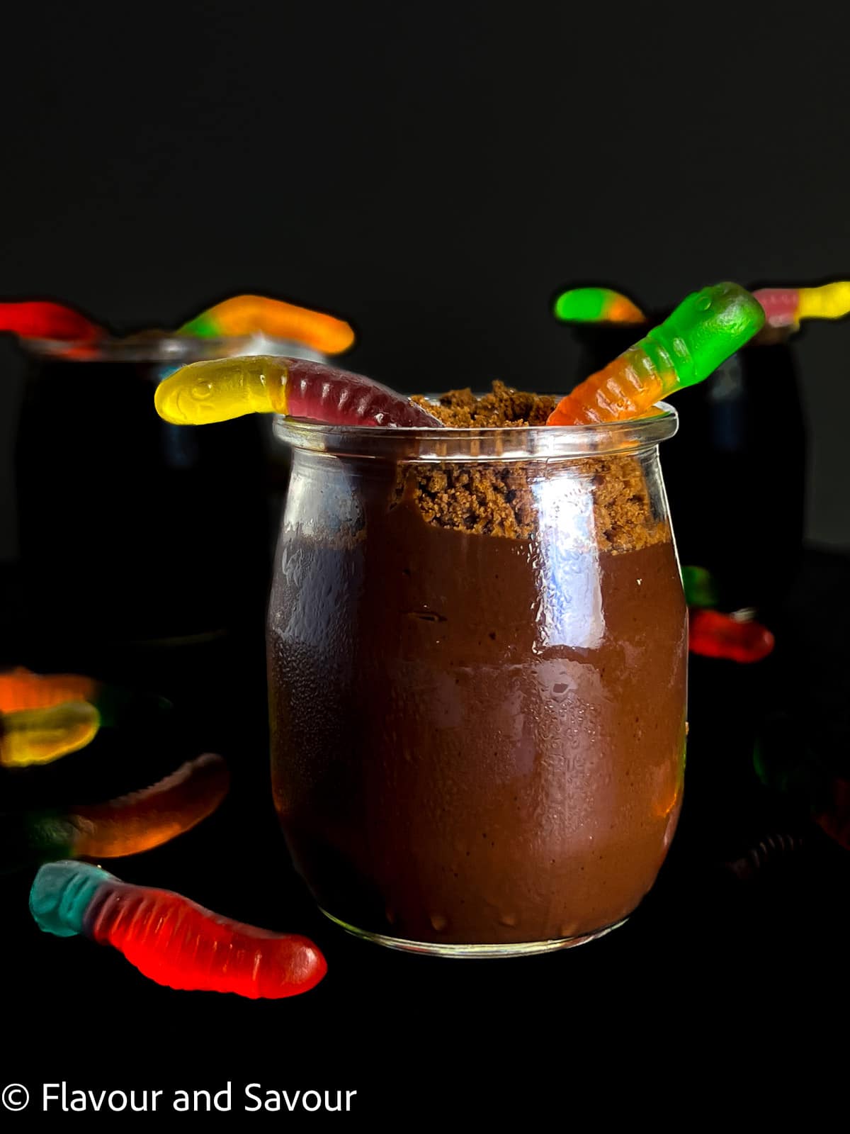A small dessert glass with chocolate pudding, chocolate cookie crumbs and colourful gummy worms.