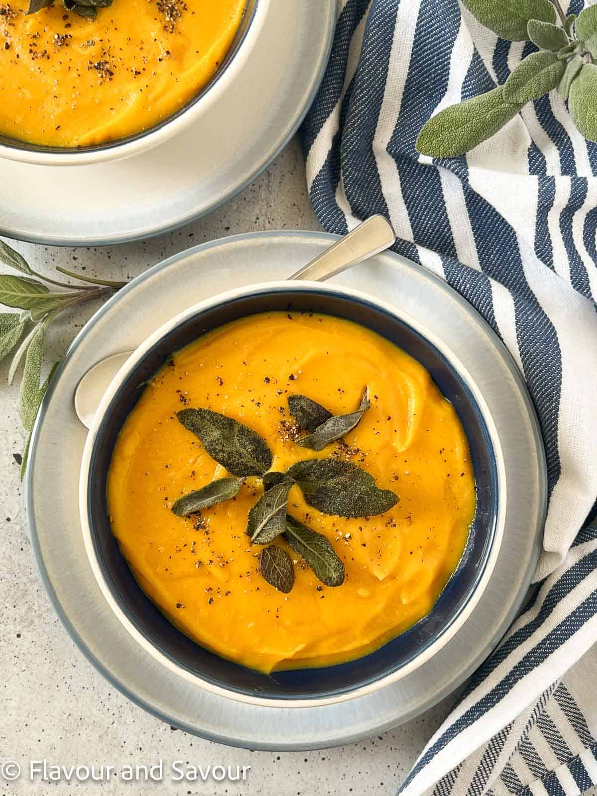 Two bowls of butternut squash soup with ginger and orange juice, garnished with toasted sage leaves.
