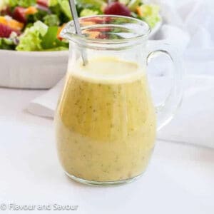 A small glass jug of everyday honey citrus salad dressing with a salad in the background.