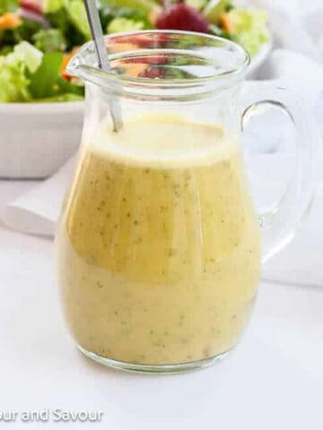 A small glass jug of everyday honey citrus salad dressing with a salad in the background.