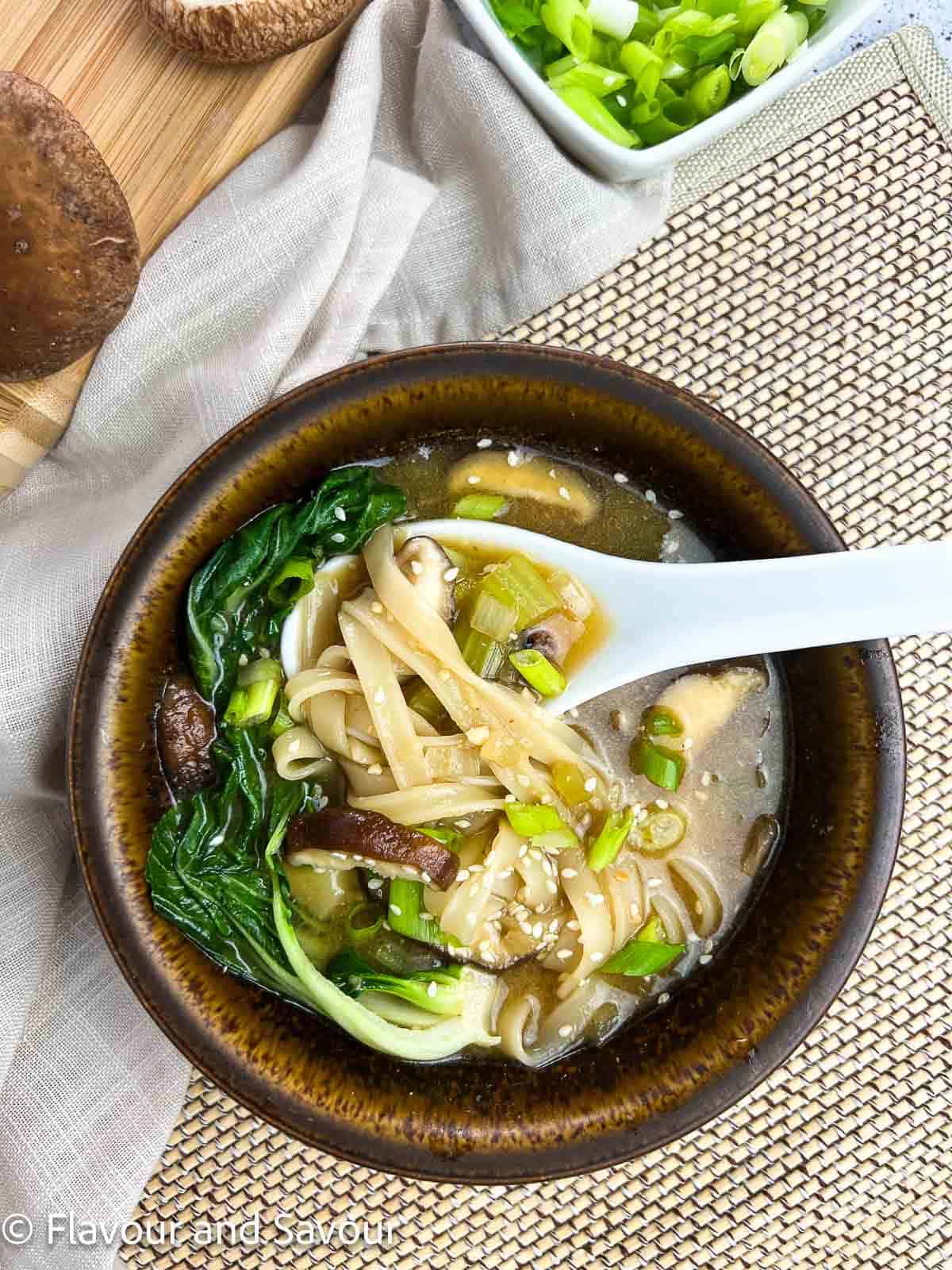 A white ceramic spoon holding rice noodles in a bowl of miso mushroom soup.