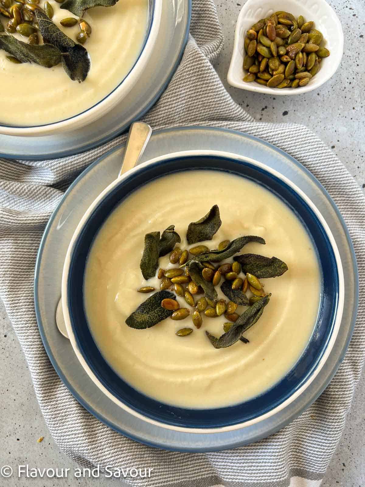 Two bowls of roasted cauliflower soup garnished with toasted sage leaves and spiced pumpkin seeds.
