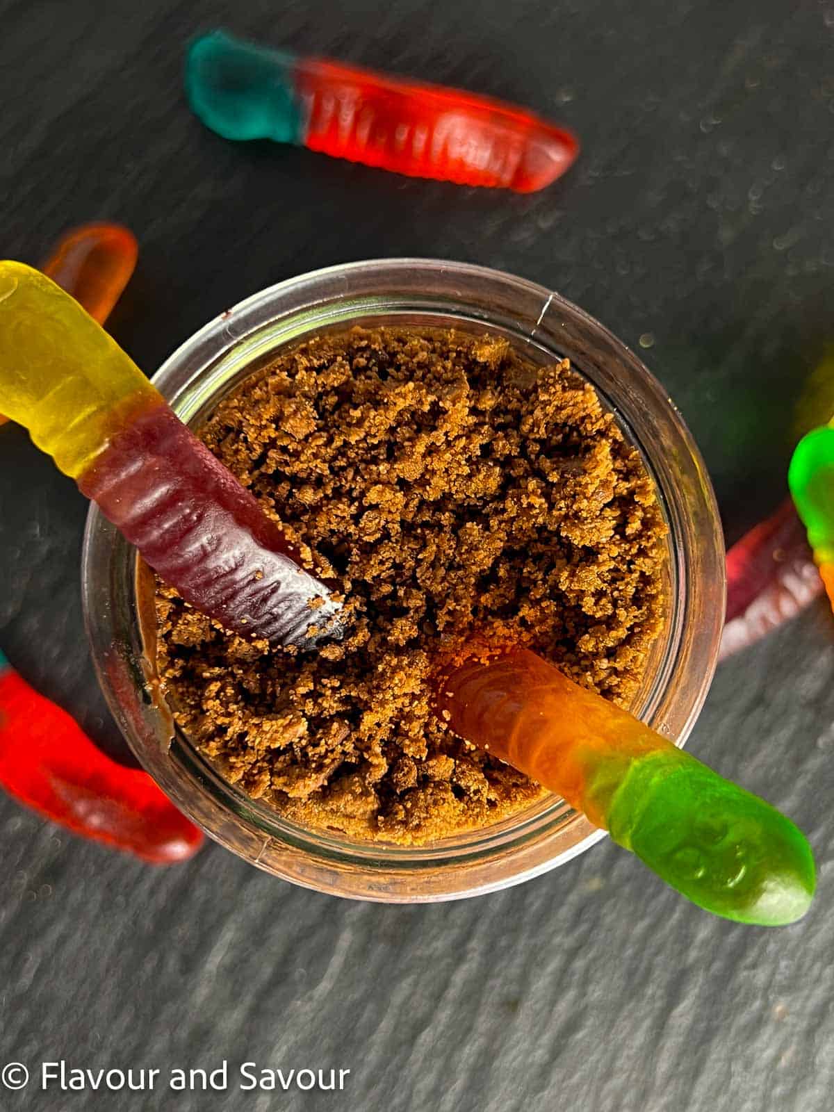 Coloured gummy worms poking out of a small dessert glass filled with chocolate pudding and topped with chocolate crumbs.