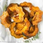 Square image of air fried acorn squash slices brushed with maple syrup, olive oil, cinnamon, sea salt and pepper with sprigs of fresh herbs beside.