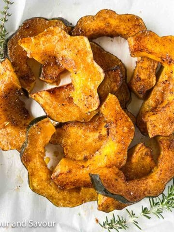 Square image of air fried acorn squash slices brushed with maple syrup, olive oil, cinnamon, sea salt and pepper with sprigs of fresh herbs beside.
