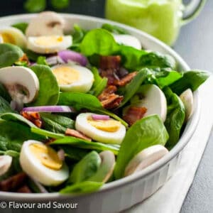 A shallow bowl with spinach, sliced hard-boiled eggs, red onion, crispy bacon and sliced mushroom.s