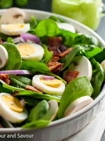 A shallow bowl with spinach, sliced hard-boiled eggs, red onion, crispy bacon and sliced mushroom.s