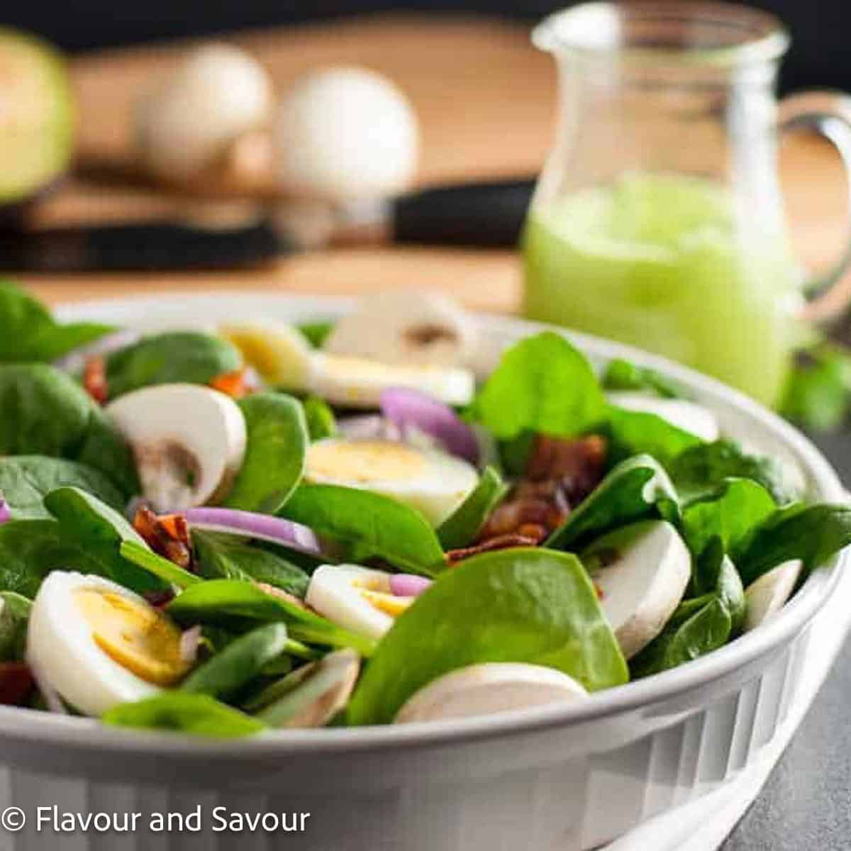 Square image of classic spinach salad with creamy avocado dressing in the background.