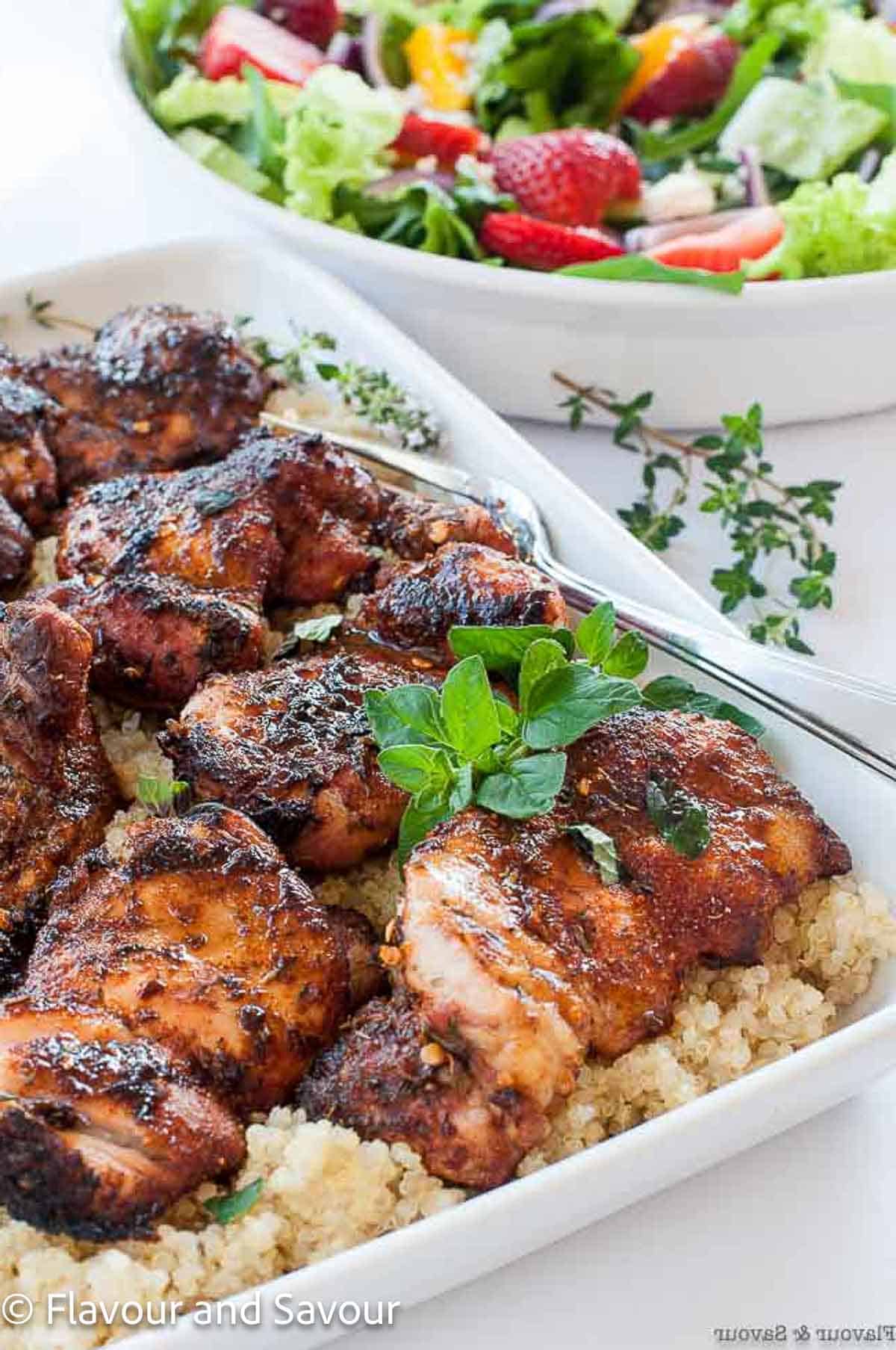 Grilled cajun chicken thighs with quinoa, garnished with fresh oregano with a colorful salad in the background.
