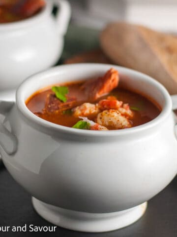Square image of a white bowl of cioppino or fish stew with a loaf of bread in the background.