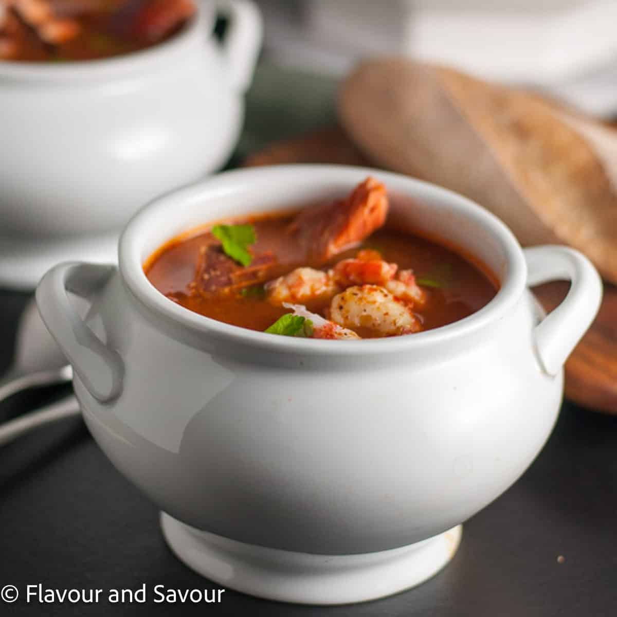 Square image of a white bowl of cioppino or fish stew with a loaf of bread in the background.