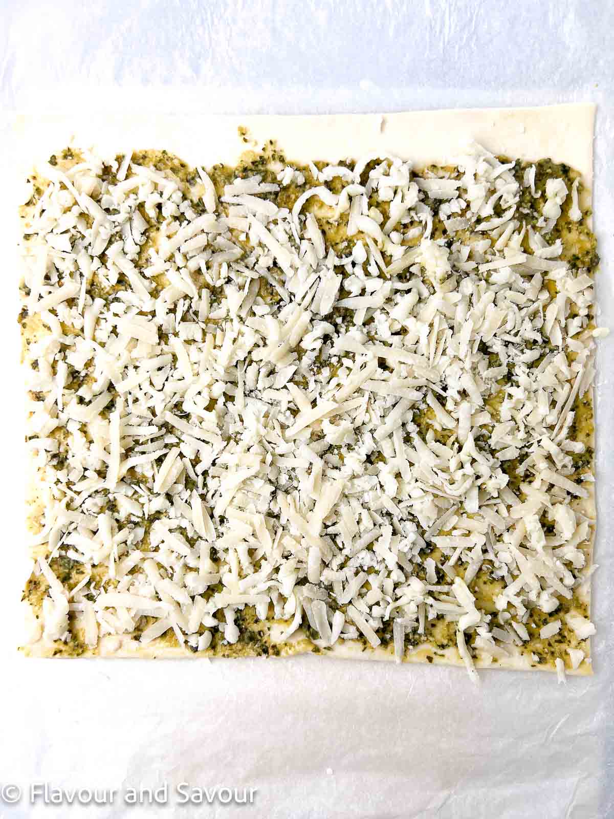 A sheet of puff pastry spread with pesto and two types of grated cheese.