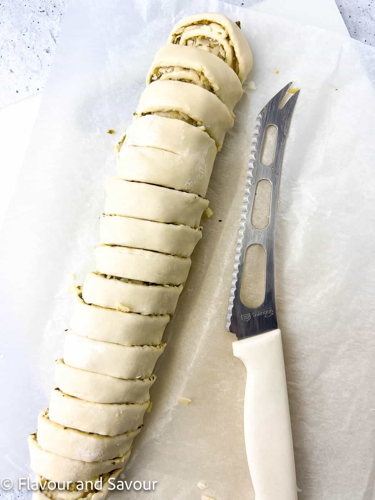 A rolled stuffed sheet of puff pastry, sliced into ½ inch slices with a knife nearby.