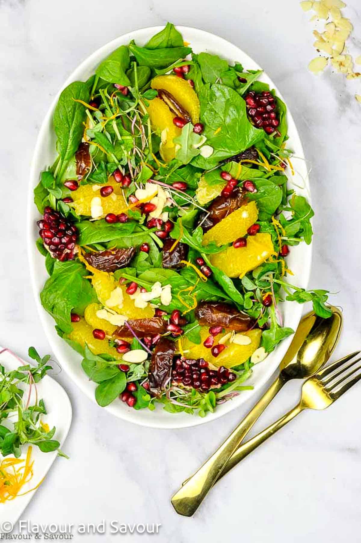 An oval white serving bowl with fresh baby spinach, arugula and oranges, dates and pomegranate arils.