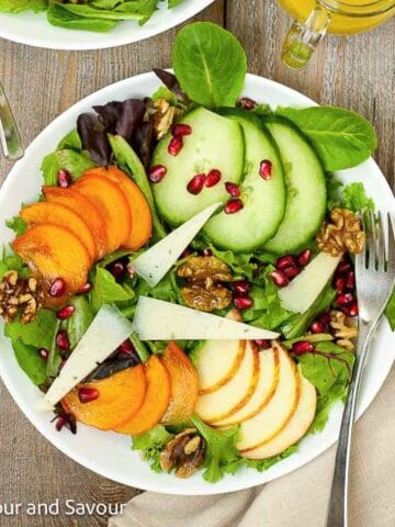 A bowl with greens, sliced persimmons, sliced apples, slice cucumbers, maple-glazed walnuts and pomegranate arils.