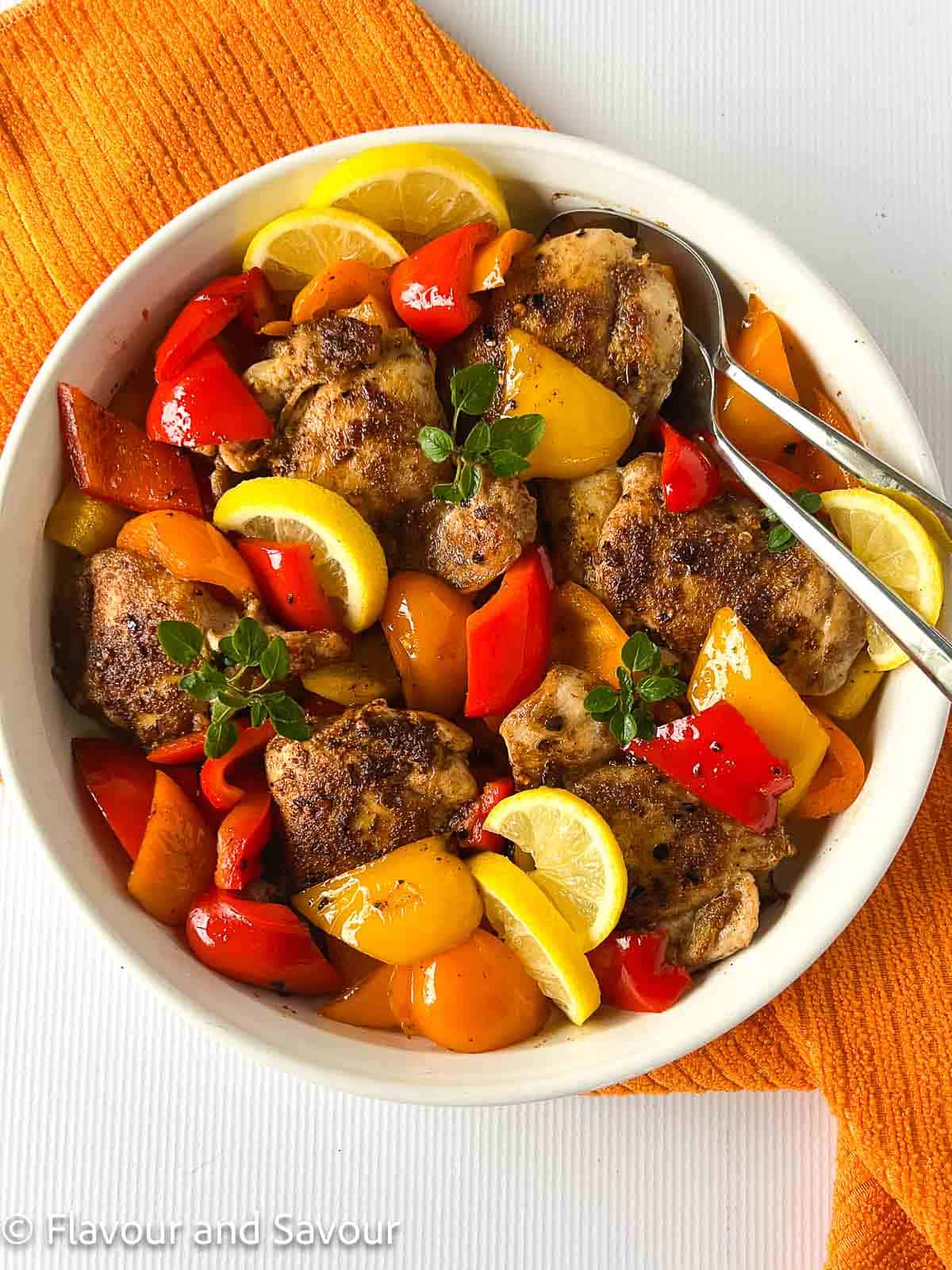 Moroccan chicken thighs and peppers in a serving bowl, garnished with lemon slices and herbs.