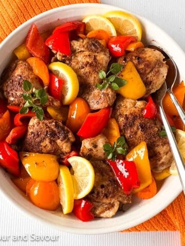 A round shallow dish with chicken thighs, colored bell peppers and lemon slices.