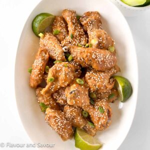 A bowl of Thai peanut chicken wings garnished with sesame seeds and lime wedges.