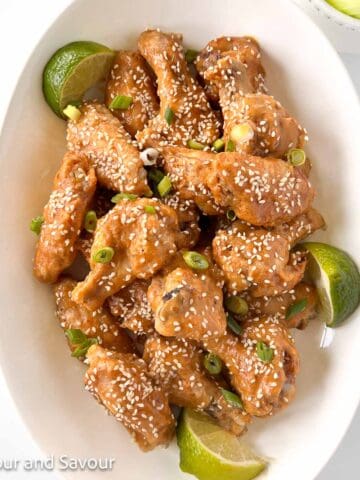 A bowl of Thai peanut chicken wings garnished with sesame seeds and lime wedges.