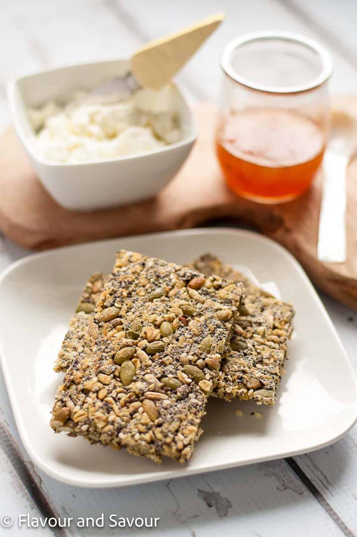 Oat crackers with seeds on a white plate with small jars of cheese and jelly in the background.