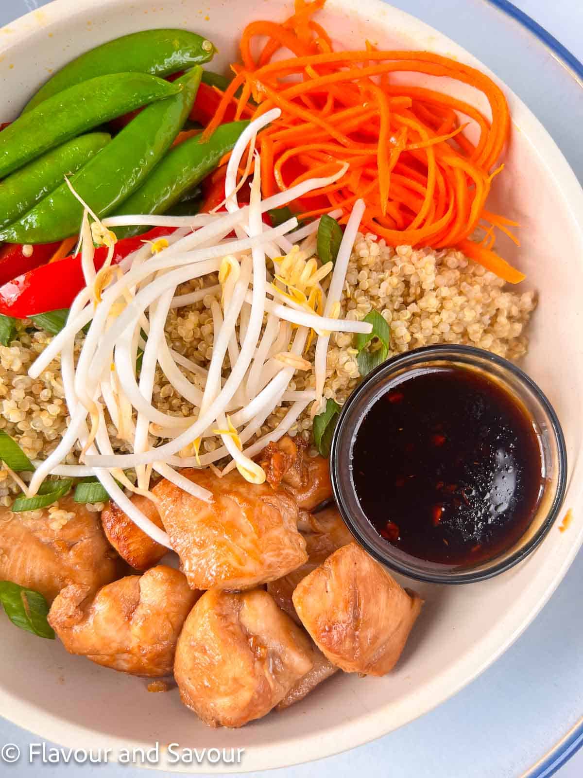 Teriyaki chicken cubes, quinoa and vegetables in a bowl with a small container of teriyaki sauce.