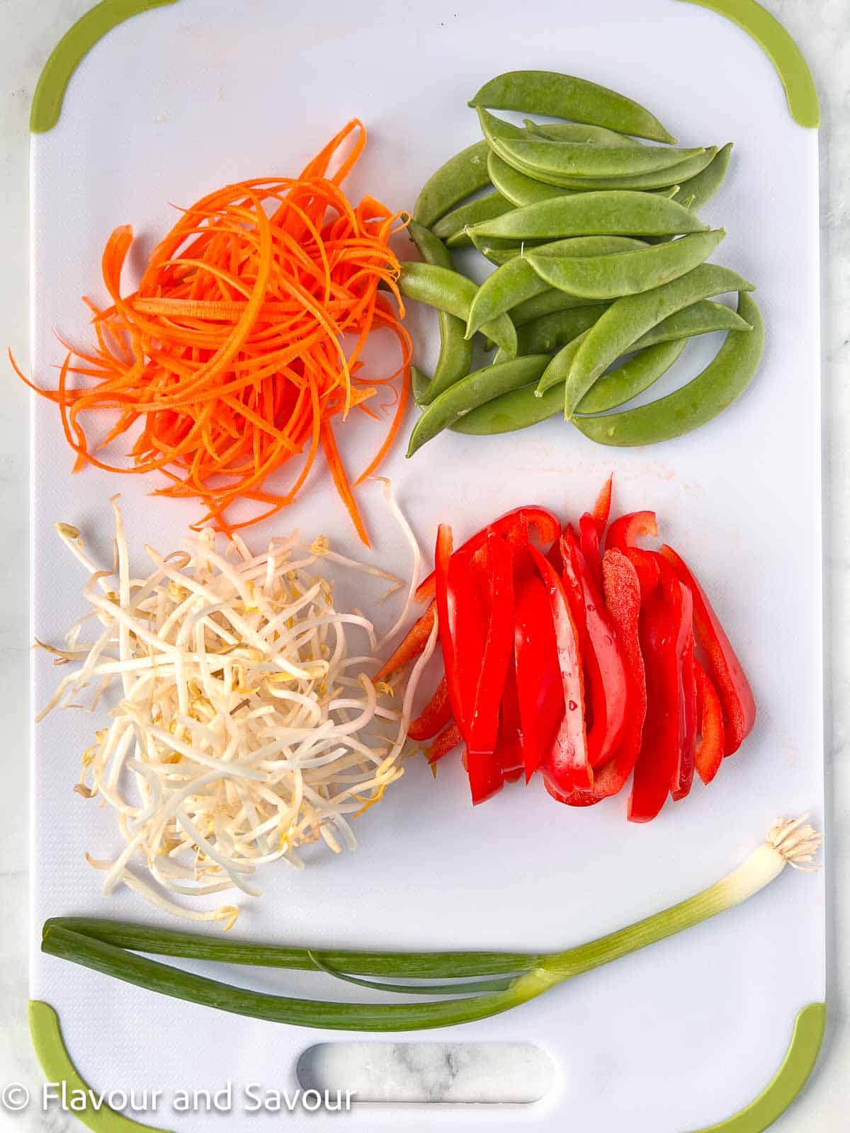 Snap peas, julienned carrots, red pepper strips, bean sprouts and a green onion on a cutting board.