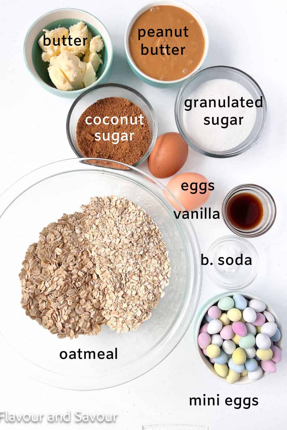 Labelled ingredients for mini egg oatmeal cookie bars: butter, peanut butter, coconut sugar, granulated sugar, eggs, vanilla, oatmeal, baking soda, candy-coated mini chocolate eggs.